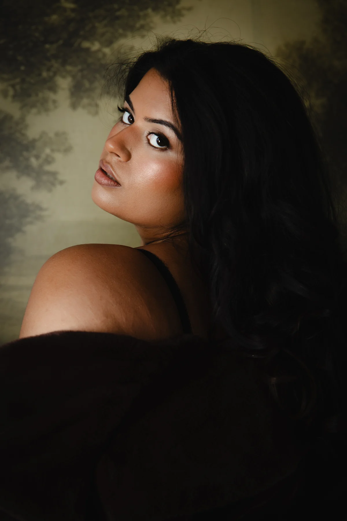 A woman looking over her shoulder, wrapped in dark fabric against a soft olive backdrop