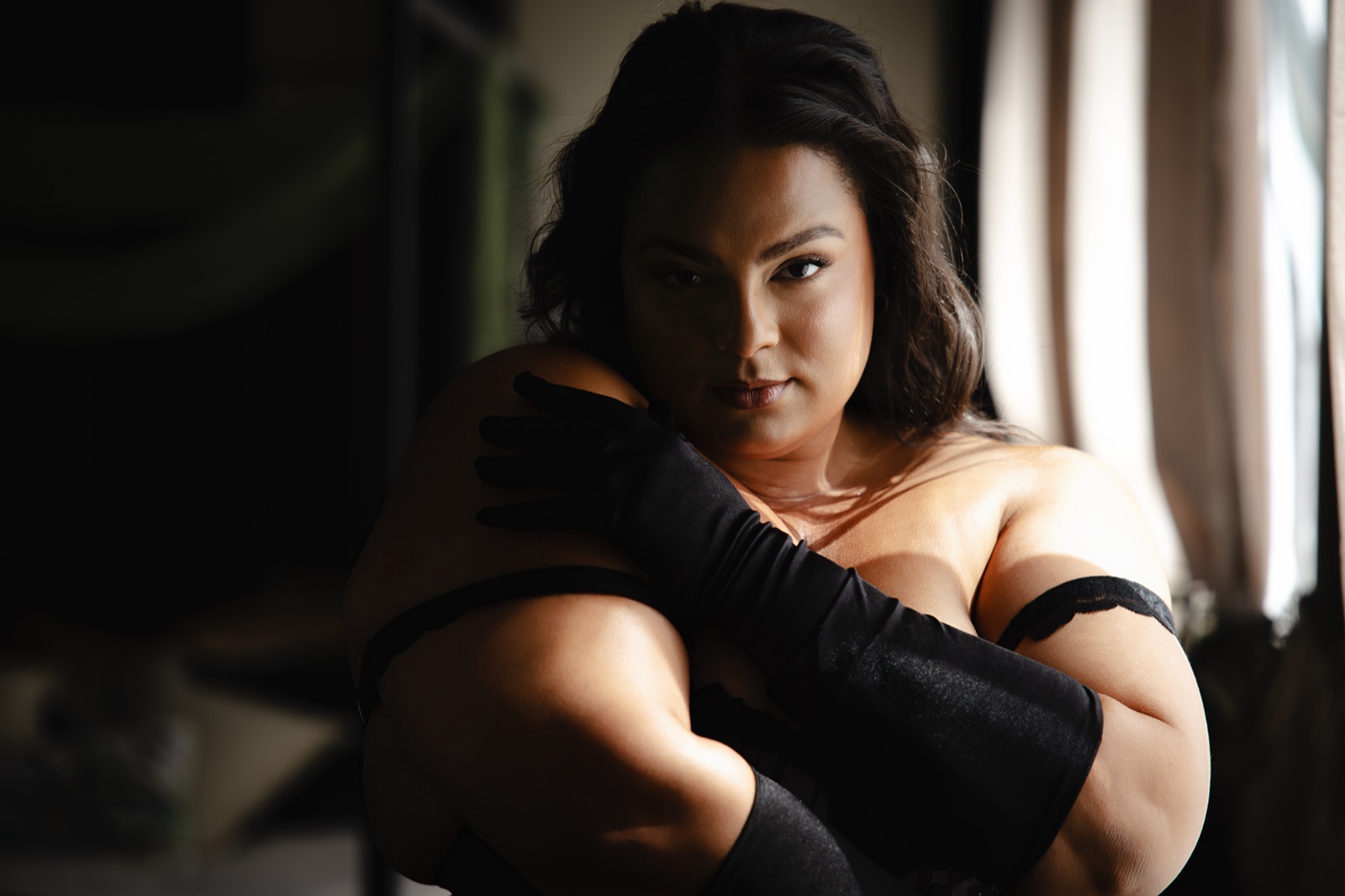 A woman in black gloves resting her hand on her shoulder in soft window light
